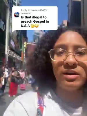 A woman with glasses and curly hair looks towards the camera. In the background, there are buildings and people on a street.