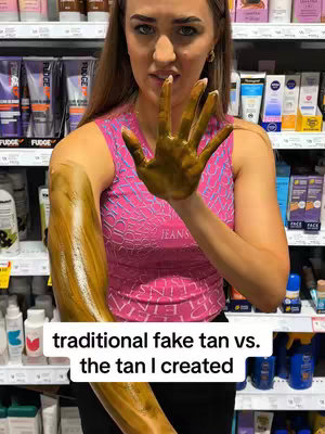 A woman is standing in a store aisle, holding out her right arm which is covered in fake tan that is a dark brown, she is showing her open hand which has fake tan on it. The rest of the arm is also covered in the same tan. The woman is wearing a pink tank top and is looking at the camera with a slight smile, her head is slightly tilted. Behind her are shelves of products, with various bottles and tubes and labels and the background is blurred.