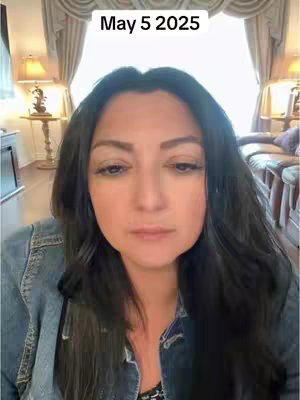 A woman is centered in a selfie-style shot. The woman has dark hair and fair skin. The background features a window with curtains, and the lighting seems soft, with warm tones. The woman gazes solemnly at the camera, with an expression that suggests thoughtfulness.