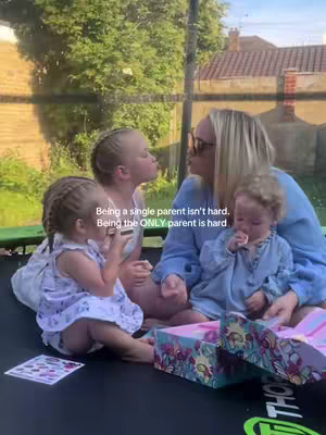 A woman sits on a trampoline with three young children. Two of the children are in front of her holding snacks, while the other child is sitting on her lap. The background shows trees and a residential building.