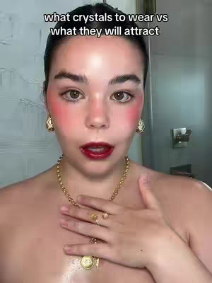 A woman with fair skin and dark hair, wears a gold necklace and earrings, with bright red blush, red lipstick, and a ring. Text overlay at the top reads, 'what crystals to wear vs what they will attract'.