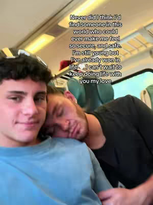 A selfie-style shot featuring two young men. One is in the foreground, looking towards the camera with a slight smile, wearing a blue shirt. The other man is in the background, resting his head on the first man's shoulder and appears to be sleeping. The setting looks like a train car, with seats and overhead fixtures visible.