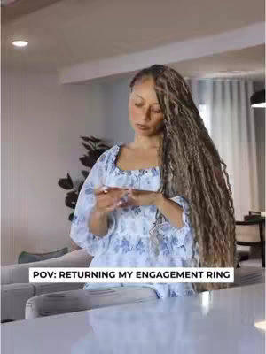 The image shows a woman in a well-lit interior. She has long, wavy braids. She is wearing a dress with a blue floral print, and her hands are near her torso as she looks down, seemingly holding or inspecting an object. The text overlay states 'POV: RETURNING MY ENGAGEMENT RING'.