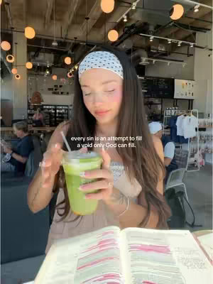 A woman with long dark hair wearing a white headband and light-colored clothing sits at a table in a cafe, holding a green drink and looking down at an open Bible. The background features cafe decor with light fixtures and other people. Text is overlaid on the image.