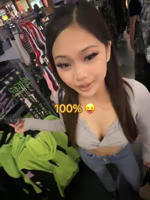 A young woman stands in what appears to be a store, holding the camera. The image is a medium shot, with the woman's face and upper body in focus. She is smiling, with her gaze directed toward the camera. She has long, dark hair, and is wearing makeup. A text overlay reads "100%" with a tongue-out emoji. The background includes clothing racks and other shoppers.