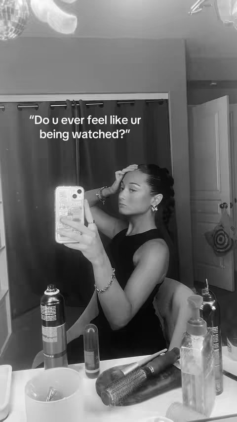 A black and white shot of a woman taking a selfie in front of a mirror. The mirror reflects a bathroom setting with various beauty products on the counter. The woman is wearing a dark top and has her hair styled back. She is holding her phone up to take the picture. The overall composition is a casual, at-home selfie.