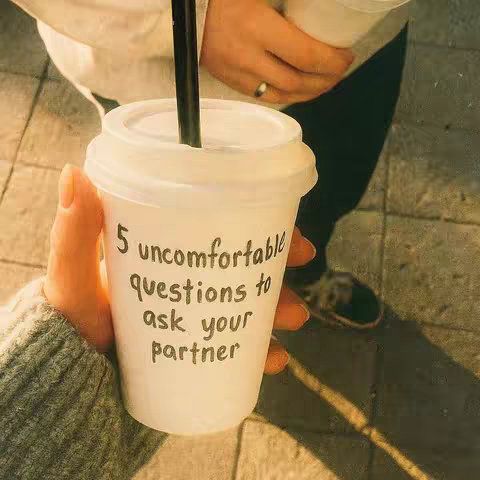 A close-up shot of a coffee cup held by a person's hand, with another person partially visible in the background. The cup features handwritten text on it.