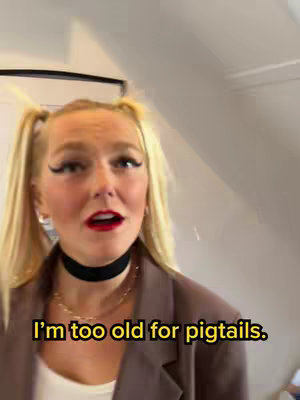 A woman with blonde pigtails, wearing a black choker and a brown blazer, is looking into the camera with a surprised expression. A text overlay reads, "I'm too old for pigtails."