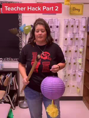 A woman stands in what appears to be a classroom holding a hammer and a purple paper lantern. The text overlay reads 'Teacher Hack Part 2'. The background includes a whiteboard, storage pockets, and a desk.
