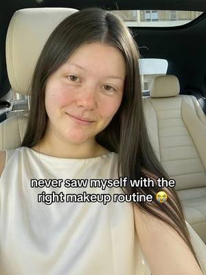 A woman smiles softly while seated inside a car. The background shows cream-colored car seats and an open window with a building visible in the distance.