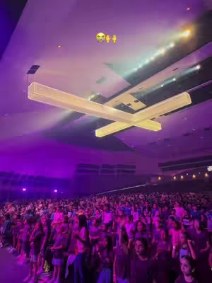 The image shows a large auditorium filled with people facing the stage, illuminated by purple and white lights. A large, modern light fixture is suspended from the ceiling. The perspective is from a low angle, looking upwards, exaggerating the size of the crowd and the hall. The overall composition is dynamic, with strong use of color and light.