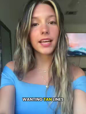 A young woman with blonde highlights in her wavy hair is centered, smiling slightly at the camera. She wears a light blue off-the-shoulder top. The background is a blurred room interior with indirect lighting. The text overlay says 'WANTING TAN LINES'.