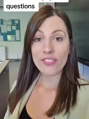 A woman with long brown hair and a beige blazer is looking directly at the camera in a classroom setting. The background includes a whiteboard and school resources.