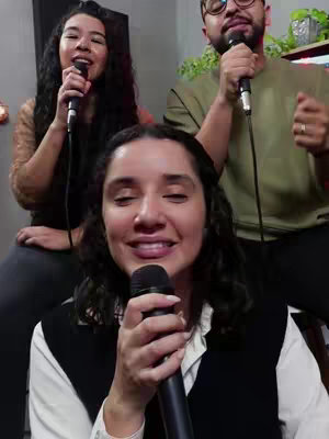 A group of three people singing into microphones. The primary focus is a woman in the foreground, smiling and holding a microphone. Behind her, there's a man on the right and another woman on the left, both also with microphones. The composition is such that the main figure is directly in front, creating a layered depth.
