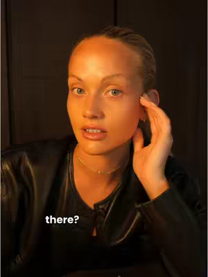 A woman with light skin and blue eyes sits in front of a dark background. She is wearing a black top and a thin gold necklace. She delicately touches her face with her right hand, her expression one of interest or questioning. Overlayed, white text reads 'there?'