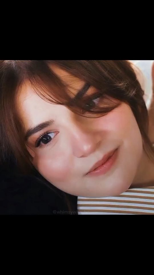A close-up shot showing a young woman's face. She's looking slightly upwards and to the side with a soft, gentle smile. Her eyes are wide and dark, and her skin has a healthy glow with blush on her cheeks. Her hair is brown, parted to the side, with some bangs framing her face. The background is blurred but seems to be a neutral light color. The overall composition is warm and inviting, and the lighting accentuates her facial features.