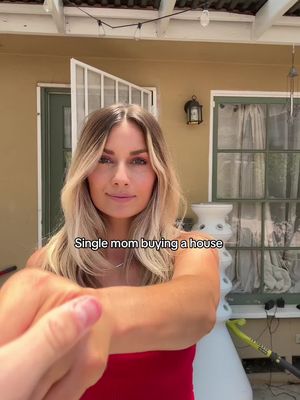 A woman with blonde hair stands in front of a house. The text overlay reads 'Single mom buying a house'. The woman is wearing a red top. A hand is holding the camera in front of the screen.