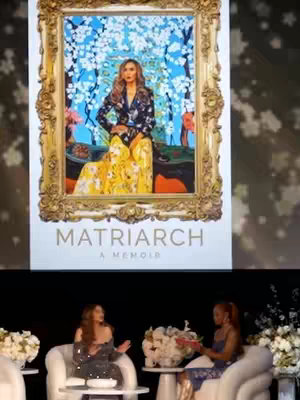 A stage setup with a large framed image of a woman, along with two women seated on white chairs with bouquets of flowers in between them. The image on the screen is framed in ornate gold.