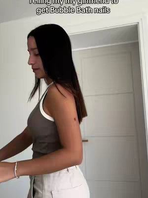 A woman stands indoors, wearing a light-colored tank top with a white trim, light-colored pants, and a bracelet on her left wrist. She is facing to one side, with dark hair falling to her shoulders, and a door and wall are in the background.
