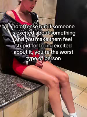 A woman sits on a stone ledge in a gym setting, dressed in a red workout outfit and black zip-up jacket. Text overlays her, conveying a relatable message about feeling judged for being excited.