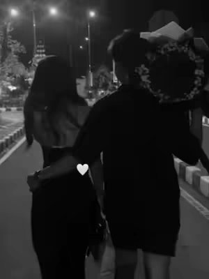 A black and white image captures a couple walking together at night. The man, carrying a bouquet, leads the way, arm-in-arm with the woman who is looking toward him. They are on a pathway. The scene is lit by streetlights.