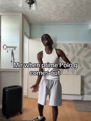 A young Black man in a white tank top and shorts is dancing in what appears to be a living room. The background includes a door, a suitcase, and a window, suggesting a casual home setting. Text overlayed on the image says, 'Me when prime Polo G comes out'.