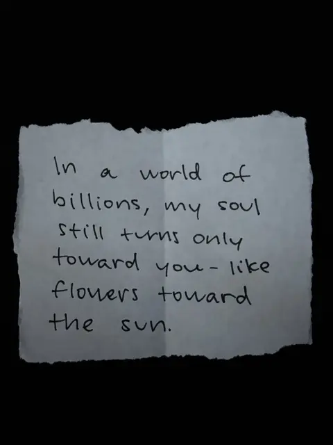 A handwritten quote is placed against a black background. The quote is written on a torn piece of paper. The paper is white and has a rough, uneven edge. The text is in black handwriting and reads: 'In a world of billions, my soul still turns only toward you - like flowers toward the sun.'