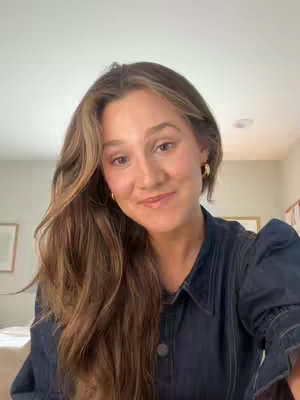 A woman looks directly at the camera with a soft smile, her wavy brown hair framing her face. She wears a denim jacket and small gold hoop earrings.