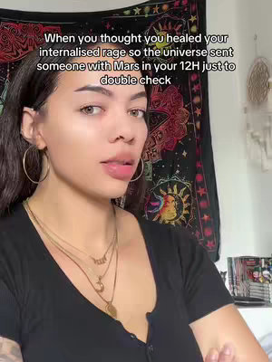 A young woman with long dark hair and large hoop earrings is featured in a medium close-up. She is looking slightly to the side with a gentle smile, her lips slightly parted. She wears several necklaces and a black top that is partially buttoned.. Behind her is a colorful tapestry and a portion of the wall.