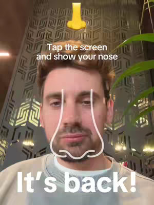 A selfie style shot of a man with short brown hair and a light colored long sleeve shirt, in the foreground in front of tall building with a complex geometric pattern. Above his head is a yellow nose graphic, and a outline of a nose over his face.