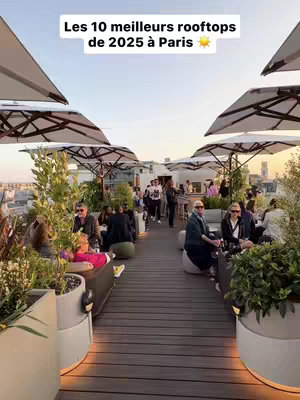 The image shows a rooftop scene with many people, some sitting and some standing, enjoying a sunny day. The composition is wide-angle, with tables and umbrellas providing shade. In the foreground, there are plant arrangements that add a touch of greenery. The overall aesthetic is bright and inviting, with a blend of natural light and artificial lighting from the lamps.