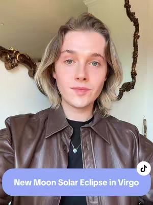 A young man with blonde hair wears a brown jacket and a black shirt, a silver necklace. The background is a light-colored wall and a decorative mirror. A text overlay at the bottom reads, "New Moon Solar Eclipse in Virgo."