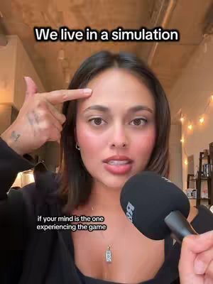 A woman with dark hair and a black jacket holds a microphone, looking towards the camera. Text at the top reads "We live in a simulation". Below, "if your mind is the one experiencing the game". Decor lights are in the background.