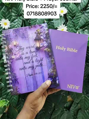 An overhead shot displays a spiral-bound journal with an illustration of a lantern and a quote, placed next to a purple book that says 'Holy Bible'. A hand is partially visible, holding the journal, set against a background of lush green leaves. Text overlays show the items' price and a phone number.