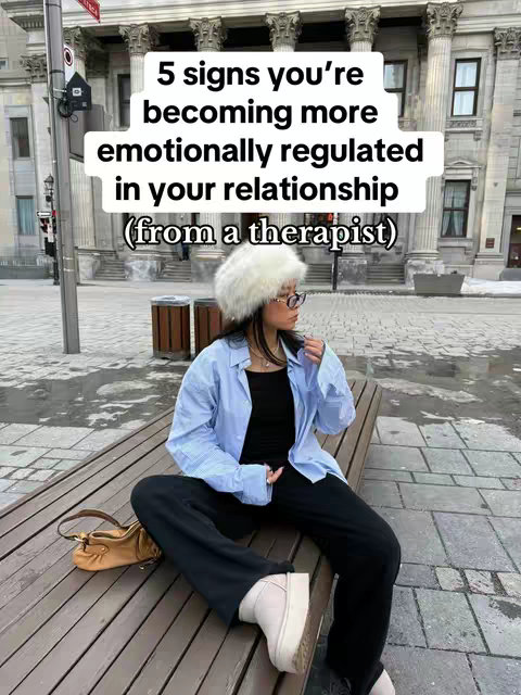 A woman wearing a white furry hat, glasses, and a blue button-down shirt sits on a wooden bench with a purse beside her. The background features a historical building with large columns, creating a city scene, and the image has overlaid text, reading '5 signs you're becoming more emotionally regulated in your relationship (from a therapist)'.