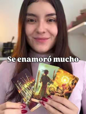 A woman with long brown hair is smiling towards the camera and holds three tarot cards. The cards are partially visible, and the background shows a blurred interior space. Text overlayed at the bottom of the image says: Se enamoró mucho.