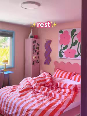 A bedroom scene with a bed covered in orange and white striped bedding, the creator is under the covers with only the top of the head visible, with a pink headboard and pillows. A pink cabinet and an abstract picture hung on the wall. Text overlayed reads 'rest' in pink.