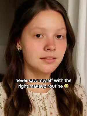 A young woman with long brown hair gazes directly at the camera in a medium shot. She wears a floral top, and the background is softly lit, suggesting interior lighting. Text overlayed on the bottom reads, "never saw myself with the right makeup routine" with a surprised emoji.