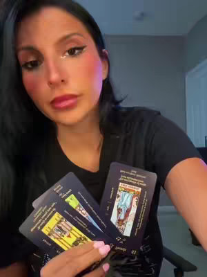 A woman with black hair is holding several tarot cards, with a soft focus and lighting. Pale pink blush is visible on her cheeks and her lips have a dark lipstick or gloss. The overall composition is intimate and focused.