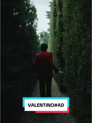 A man in a red suit walks away from the camera down a narrow pathway defined by tall green hedges.
