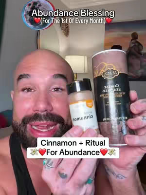 A bald man with a beard holds up a jar of cinnamon and another container. The background shows what appears to be some kind of artwork with a lamp and a fan. Text overlays with a moon, heart emojis and dollar signs.