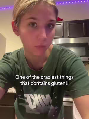 A woman with short blonde hair is looking directly at the camera with a neutral expression. The background shows a kitchen with dark cabinets and stainless steel appliances, and the top of the frame has purple string lights.