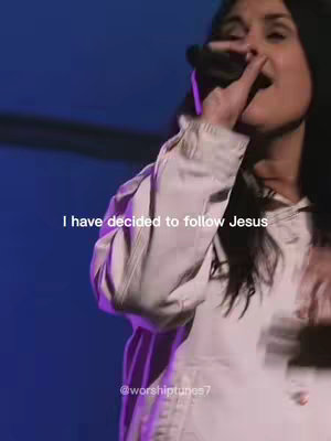 The image is a vertical video still showing a woman on a stage or in front of a blue backdrop with soft lighting. The woman is singing into a microphone, with her right hand near her mouth. Overlayed text says 'I have decided to follow Jesus'. The image has a slightly desaturated look.