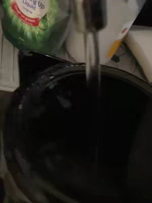 The image captures a close-up view of a sink with the faucet running, pouring water into a dark basin. Partially visible are a green bottle of 'Liquid' dish soap, placed near the faucet.