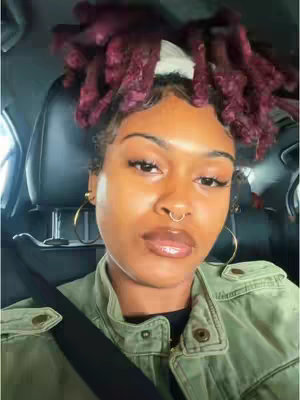 A close-up shot of a person in a car, showing the person's face. The person's hair is styled in maroon dreadlocks that extend upwards from the head. The person has dark skin and a forward gaze. The person wears a light green jacket and gold hoop earrings. The person has a nose ring. The background is blurred but suggests the interior of a vehicle.