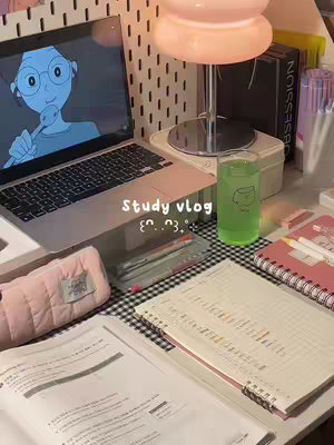 A well-lit top-down photograph showcases a tidy study setup. The central focus is a laptop displaying a cartoon animation. Surrounding the laptop are school supplies like notebooks, pens, and an open textbook, complemented by a green-tinted drink and a stylish pink lamp. The setting is clean and organized, giving a sense of a productive study environment.