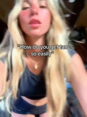 A selfie-style shot of a young woman with long, blonde hair. She is looking directly at the camera with her mouth slightly open. The overlay text, in a white box, asks, "How do you get tan so easily"
