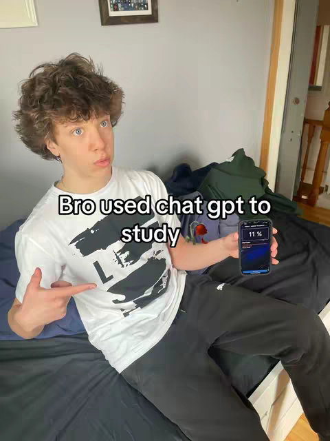A young man with curly brown hair points towards a smartphone in his hand, displaying an app that says 11%. The man is wearing a white t-shirt and black pants, sitting on a bed. The background includes a wall, a picture frame, and a glimpse of a wooden structure.