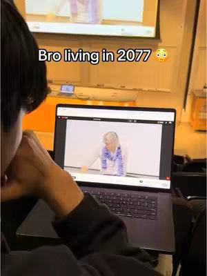 A person is shown from the chest up, they are seated with a laptop in front of them, displaying a youtube video. There is a large screen in the background, with some light fixtures showing.