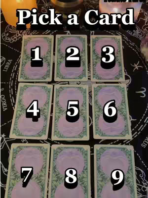 The image displays a tarot reading setup. Nine tarot cards are laid out face down with numbers 1 through 9 in the center of each card, and the words "Pick a Card" are displayed at the top. The cards are on a black surface with astrology symbols visible on the border. Two candles and other items are also visible in the image.